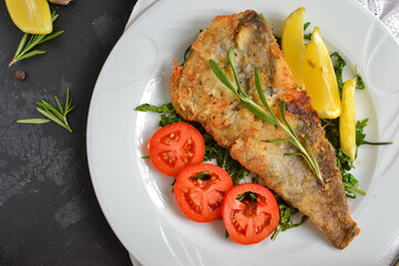 Fried fish fillet with tomatoes, lemon and rosemary. White plate. Dark background. White linen napkin. Top view. Closeup