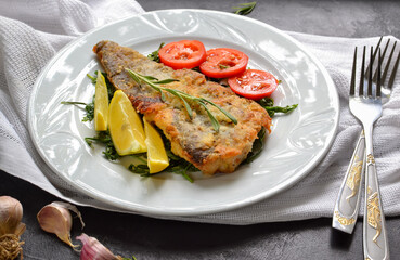 Fried fish fillet with tomatoes, lemon and rosemary. White plate. Dark background. White linen napkin.