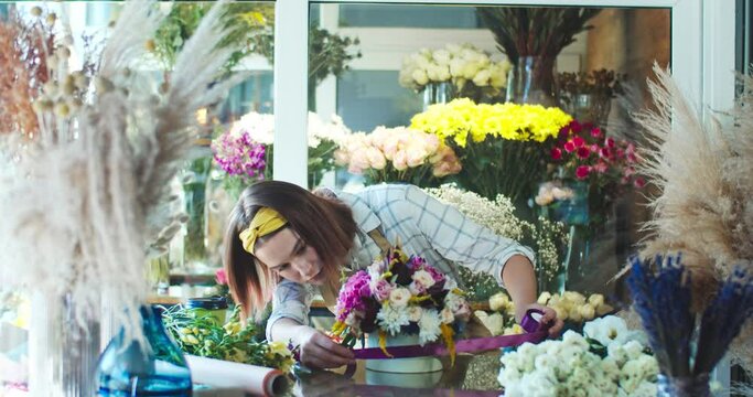 Pretty Caucasian woman wearing apron, working in flower store. Good-looking young female florist is tying purple ribbon around hatbox while decorating blossom bouquet. Decor, business concept.
