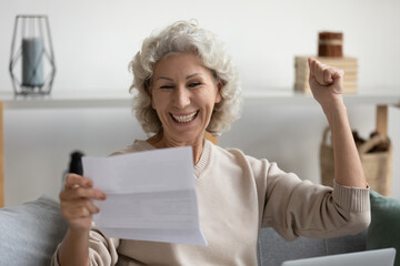 Overjoyed smiling middle aged woman reading good news in letter close up, showing yes gesture,...
