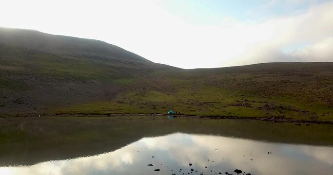 Fly above a beautiful stunning calm quiet lake in mountain highlands a couple and kids camping setup a blue travel tent by the coast in a sunny cloudy day in Iran
