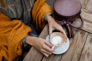 Woman drinking coffee . Stylish bag on table. Wearing grey dress and  orange plaid. Enjoying cozy morning in cafe.