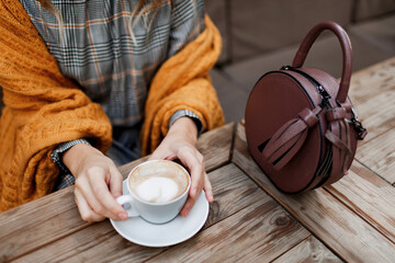 Woman drinking coffee . Stylish bag on table. Wearing grey dress and  orange plaid. Enjoying cozy morning in cafe.