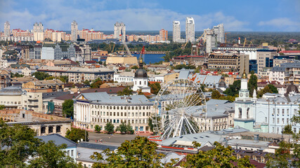 The landscape of the summer city of Kyiv overlooking the old district of Podil with a Ferris wheel and a bell tower with a gilded dome, the Dnipro River and many city buildings.