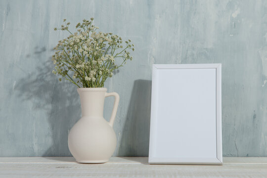 A vase with flowers and a white photo frame on the table. Light stylish background. Preparation for inscriptions, copy space.
