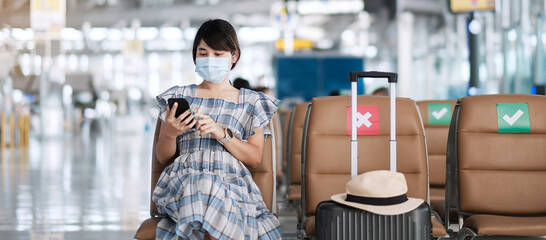 Young female wearing face mask and using mobile smartphone in airport, protection Coronavirus disease (Covid-19) infection, Asian woman traveler sitting on chair. New Normal and social distancing