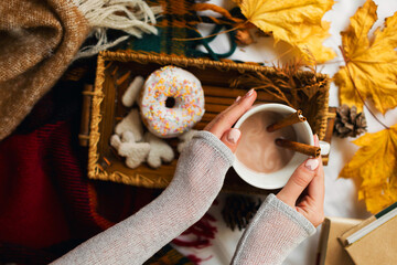 Lifestyle image of pretty girl eating tasty breakfast in bed on wooden tray with cup of cacao, cinnamon, cookies and glazed donuts. Wearing warm sweater and woolen socks.
