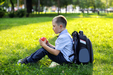 charming blond boy 6 years old in a light shirt and bow tie with a backpack on his back sits on the green grass in the Park and eats an Apple