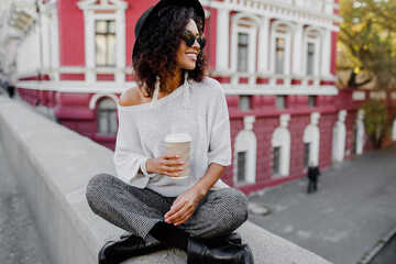 Street fashion look. Stylish black girl sitting on the bridge and holding cup of coffee or tea...