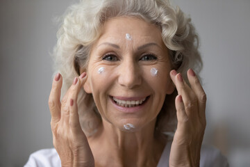 Head shot portrait smiling beautiful mature senior woman applying moisturizing face cream,...
