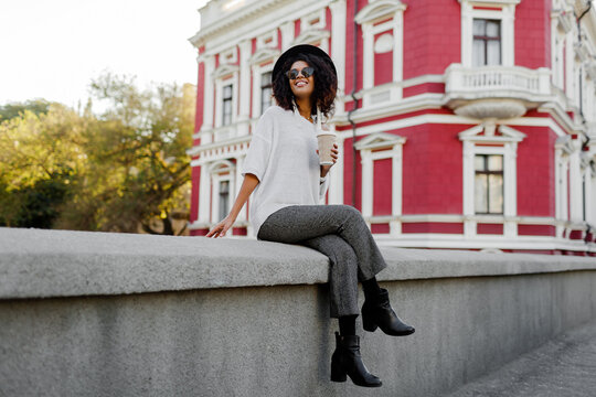 Full height image of Playful black woman with Afro hairs sitting on the bridge and having fun. Wearing leather boots and whoop  trendy trousers Happy leisure time in old European city.