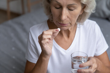 Close up mature woman holding white round pill and glass of water, sitting on bed at home, unhealthy middle aged female taking painkiller medication, antibiotic or supplement, relieving pain