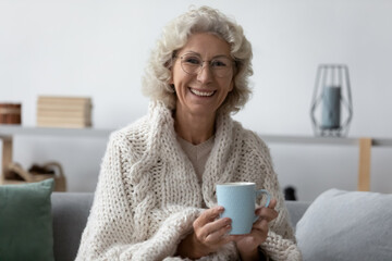 Head shot portrait smiling mature woman wrapped warm blanket holding mug of tea or coffee, happy...