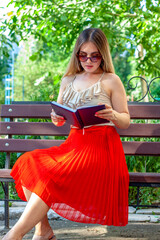 One beautiful girl in a dark sunglasses, red skirt and white top sits on the bench outside  and...