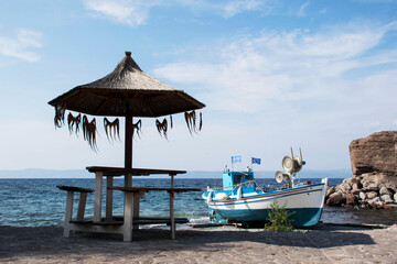 Fototapeta premium little harbor of greek village on lesbos island. One small fishing boat. Sea on the background. Summer season. Drying squid hanging beneath beach umbrella. Fishing. Sea food. Calamari hanging to dry.
