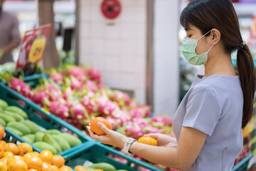 Asian woman wearing surgical face mask and holding orange fruit in supermarket or grocery, protect coronavirus inflection. Hygiene, new normal and life under covid-19 pandemic