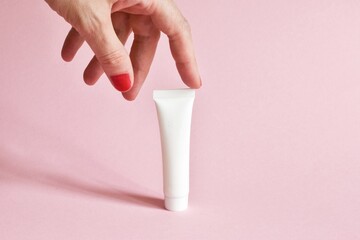 Female hand holding white cosmetic product tube. Blank plastic container for cream, lotion, toothpaste, nourishing or moisturizing mask on a pink background. Eco-friendly, organic cosmetology concept.