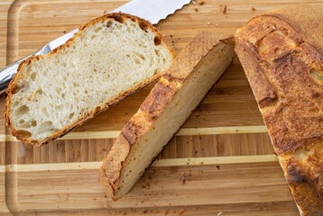 Food composition in natural light. Crispy homemade bread (French roll), sliced on a wooden board. Nearby is a fragment of a metal knife