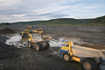 Obraz premium Excavation in a mountainous area, dump trucks queue up to be loaded in a mining quarry. A bulldozer is working nearby.