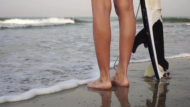 A woman with a surfboard stands on the seashore, legs and waves close-up