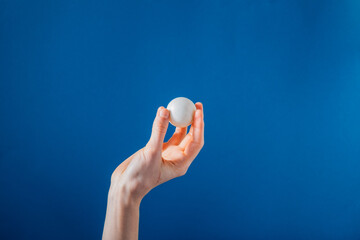 Hand Holding Ping Pong Ball On Blue Background Photo A hand holds up a white ping ping ball in front of a bright blue background.
