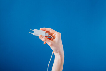 Holding A White Phone Charger On Blue Photo A hand holds up a white European phone charger, the...