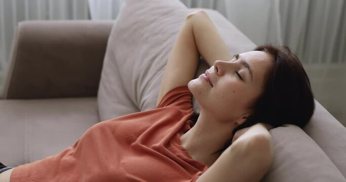 Close up serene woman puts hands behind head relax leaned on sofa cushion in living room, enjoy calm time at home, breath fresh humidified air at summer day, no stress, fatigue relief, harmony concept