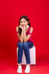 Portrait pretty young asian woman. Lovely girl sitting on a chair. She wear jeans, snickers shoe. Cute female is good looking girl, she has nice smile. red background, copy space, taken full length