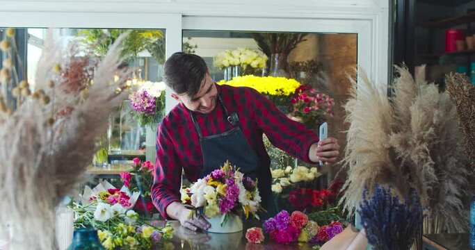 Good-looking young male florist working in modern flower store. Charming Caucasian man calling customer to show bouquet in shop. Business, commerce, entreprenuership concept.