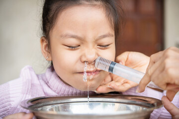 Little asian girl saline nasal wash nose at home. Asia mother making nasal wash for her kid girl by flushing nose cleaning with syringe and saline to treat the flu. health care medical people concept. - obrazy, fototapety, plakaty