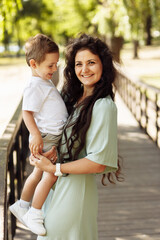 Fototapeta premium Portrait of adorable mother with overjoyed son at the park, beautiful woman hold in arms little boy, smiling, family time, motherhood and childhood concept