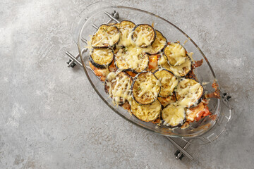 Gratin from eggplant, tomato and bell pepper with cheese, baked in a glass casserole dish on a gray concrete background, copy space, high angle view from above
