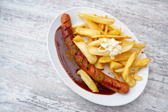 Currywurst, popular fast food in Germany consisting of sausage with curry ketchup, served with French fries and mayonnaise on an oval plate on a grey background, copy space, high angle view from above