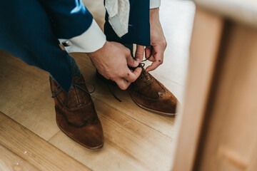 A man ties up his shoelaces on his brown wedding shoes in the room. Blue suit and patent leather...