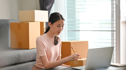Beautiful girl writing costumer address on box while sitting at the modern living room. SME business startup.
