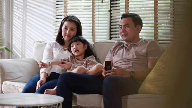 Happy asian family bonding laughing watching tv sit on sofa in modern new home, cheerful loving young parents embracing little cute kids cuddling on couch.