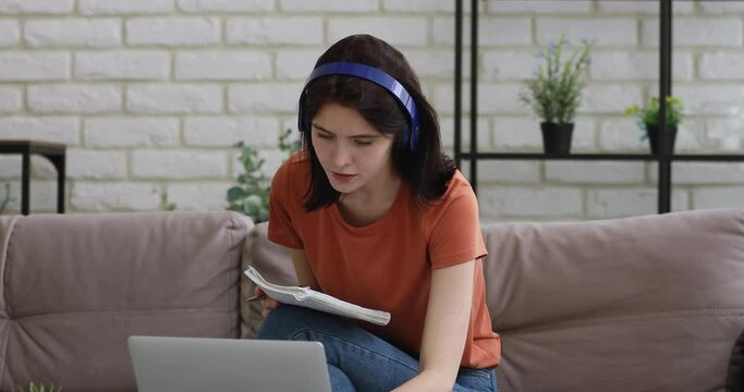 Girl wear headphones sit on couch writes notes in copybook discuss common task with mates using laptop and video conference application, easy convenient e-learning process modern tech usage concept