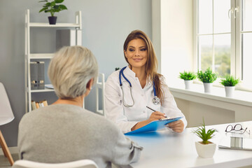 Fototapeta premium Smiling woman doctor listening to senior woman patient and making notes in medical clinic