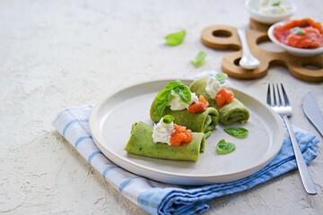 Spinach pancakes rolled up on a white plate on a light concrete background. Served with salted salmon and soft cheese.