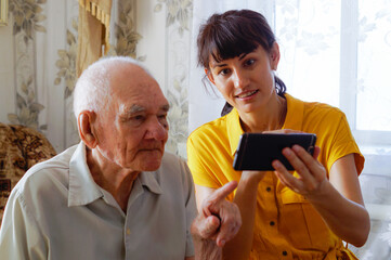 a man of the 90s with an adult granddaughter, sitting together on a cozy sofa with a phone in his...