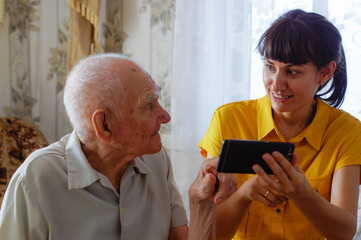 An old grandfather and an adult granddaughter look at photos on their phone. A young woman shows an...