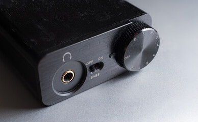 Close up shot of a small digital audio converter, or DAC, on a silver metallic surface.  Volume dial, headphone jack, and bass boost switch.  Black metal device for audiophiles.