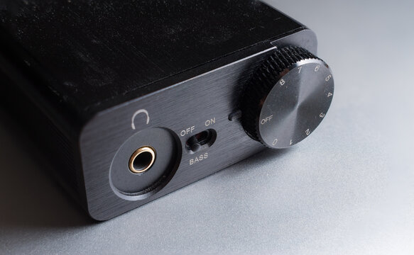 Close Up Shot Of A Small Digital Audio Converter, Or DAC, On A Silver Metallic Surface.  Volume Dial, Headphone Jack, And Bass Boost Switch.  Black Metal Device For Audiophiles.