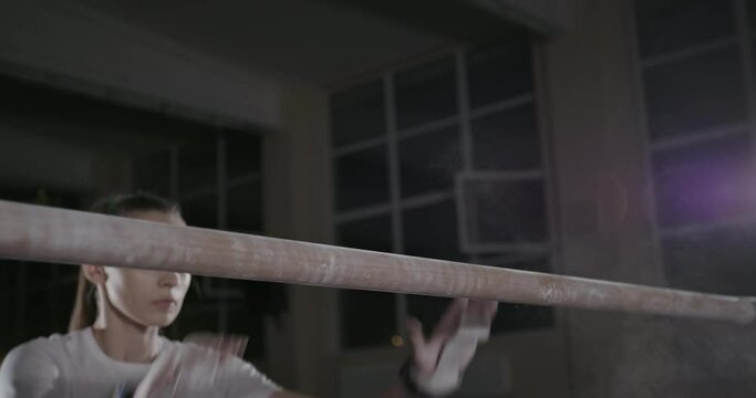 Tilt Up View Of Young Female Gymnast With Chalk On Hands Turning On Bar During Training In Dark Gym