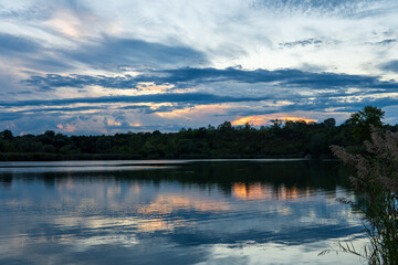 Orange striking sunset above the lake with sky, bright clouds and green forest trees reflecting on the water
