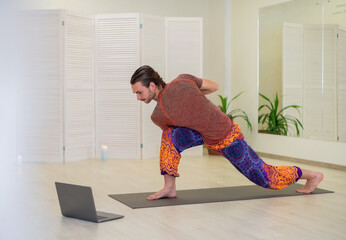 A young male yogi practices and teaches yoga online on a laptop in the Studio, performing and...