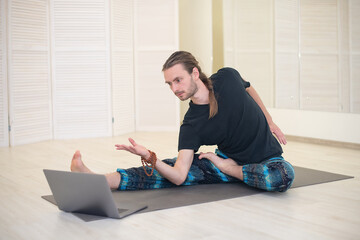 A young male yogi practices and teaches yoga online on a laptop in the Studio, performing and...