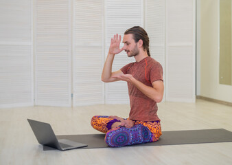 A young male yogi practices and teaches yoga online on a laptop in the Studio, performing and...