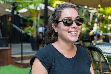 Young woman wearing sunglasses, smiling, waiting, sitting in an outdoor bar.