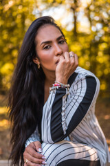 Summer lifestyle, young Latin brunette girl in a very elegant white suit with black stripes. In a beautiful park sitting, portrait looking at camera